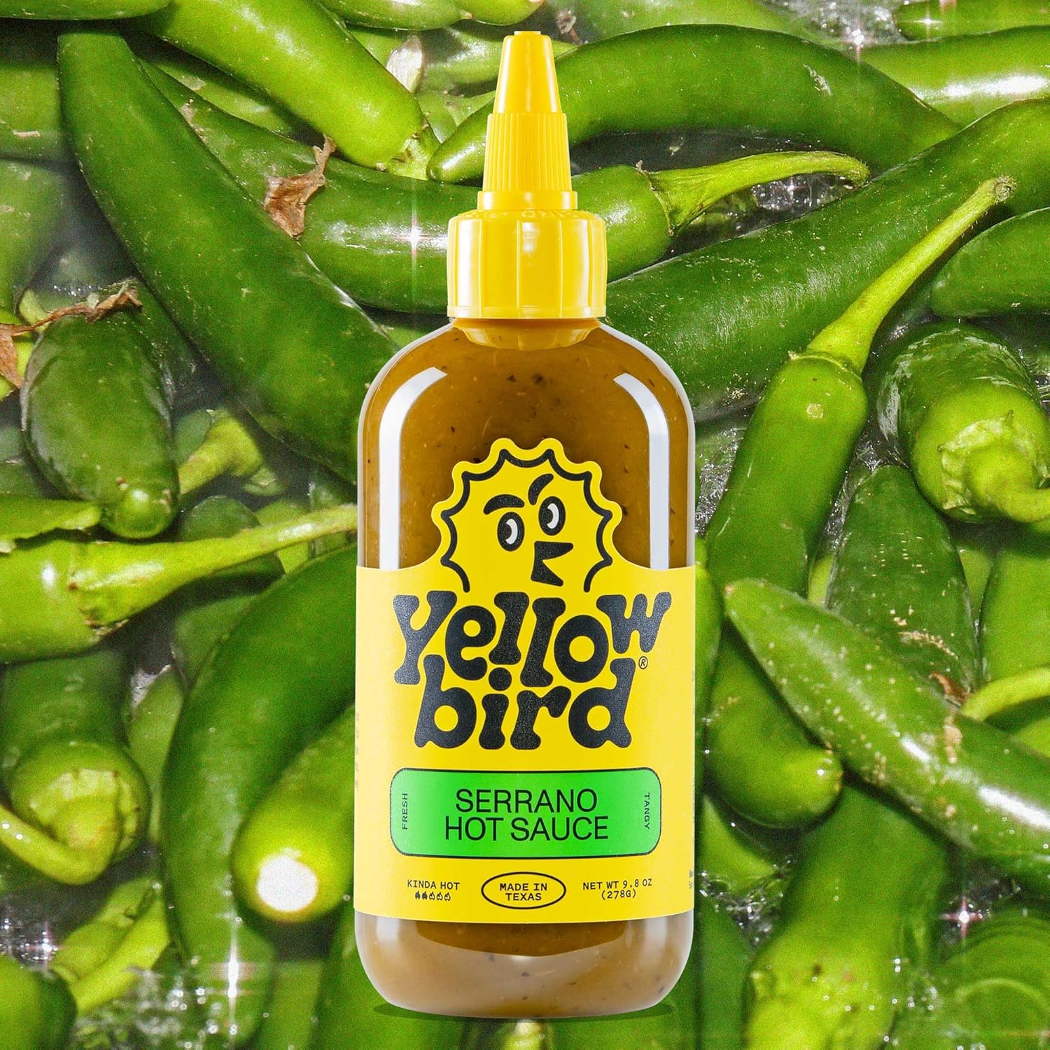 Yellow bird Serrano Hot Sauce bottle surrounded by green peppers