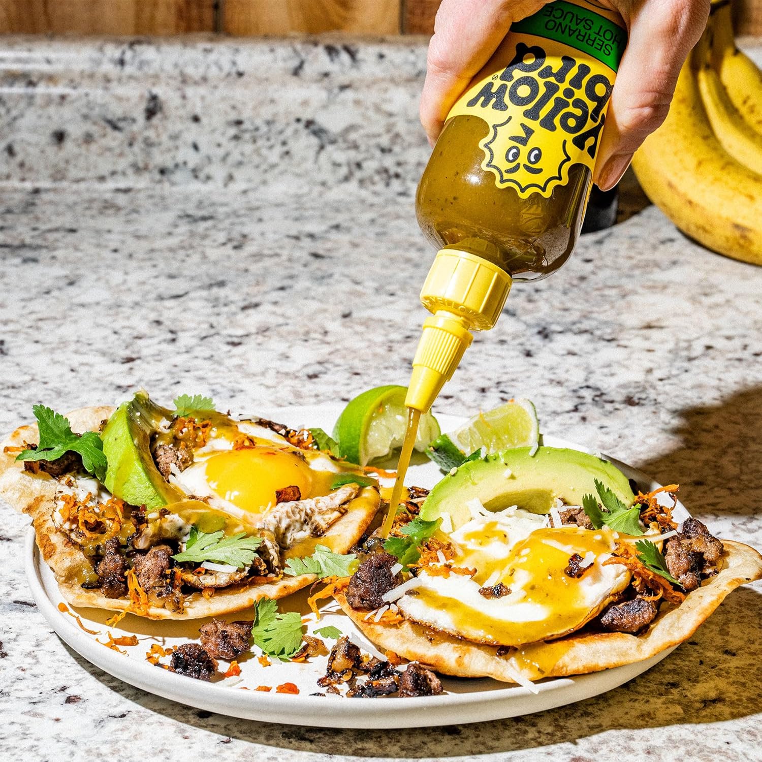 Person pouring Yellowbird Serrano hot sauce over a dish with eggs, avocado, and other toppings on a marble surface.